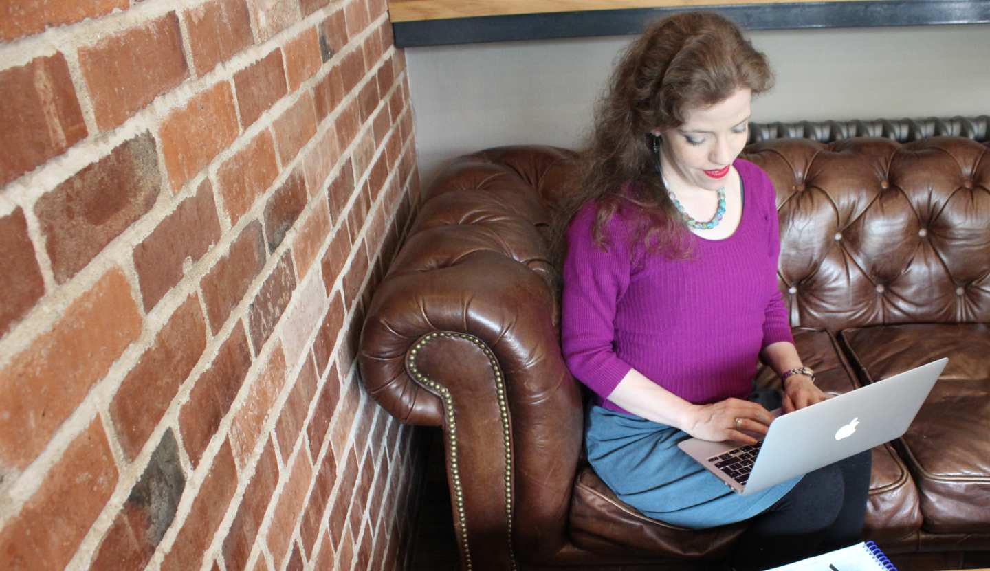 Grace sitting working on a silver macbook, wearing a purple jumper and teal skirt. 